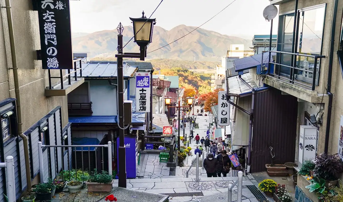 Fotografía real de la escalinata de Ikaho Onsen en Gunma, Japón, con tiendas tradicionales y montañas al fondo, localización que inspiró Dandadan